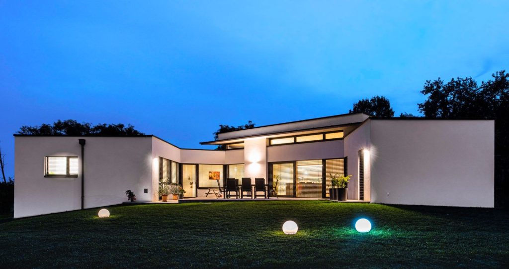 Moderne Bungalow-Villa in L-Form bei Nacht. Weiße Fassade mit großen Fensterfronten, die warm erleuchtet sind. Im Vorgarten befinden sich drei beleuchtete Kugelleuchten auf dem Rasen. Der Himmel ist in der blauen Stunde zu sehen.